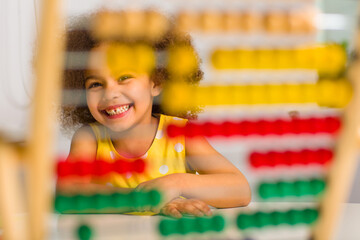 A Black student in a yellow dress laughs brightly behind a colored abacus in an elementary school