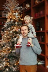 Smiling Caucasian father holding baby girl on shoulders by decorated Christmas tree. Happy family celebrating Christmas or New Year winter holiday at home. Winter holidays mood, spirit.