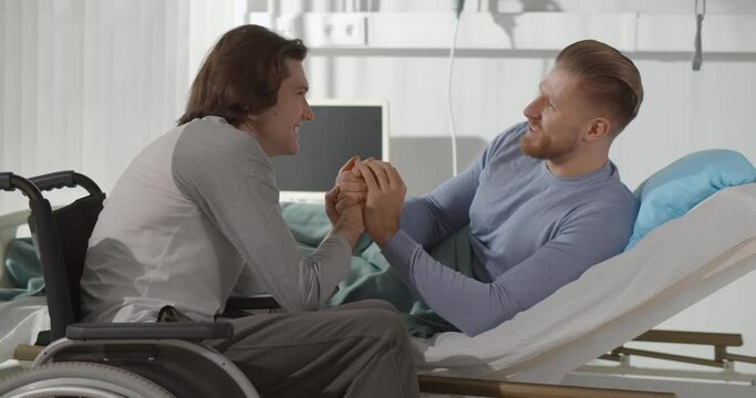 Disabled young man in wheelchair holding hands of sick boyfriend lying in hospital bed