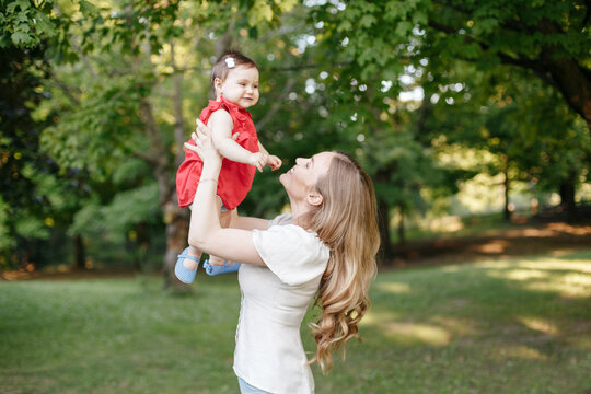 Happy Childhood. Young Smiling Caucasian Mother And Girl Toddler Daughter Playing In Park. Mom Tossing Throwing Child Baby In Hands On Summer Day Outdoor. Authentic Family Childhood Lifestyle.