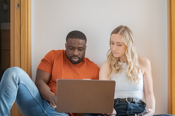 Young interracial couple sitting on the floor while using the laptop and consulting the mobile