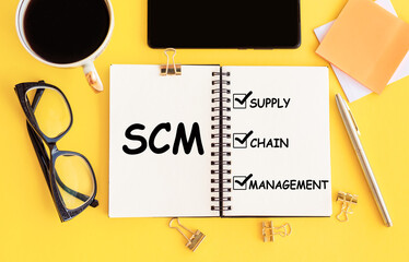 SCM - Supply Chain Management acronym, on notepad and office accessories on yellow desk.