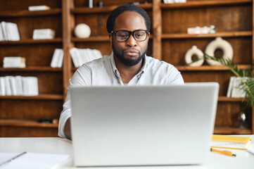 Concentrated African-American man wearing stylish eyeglasses and smart casual shirt using a laptop for work sitting in the modern office space, a multiracial office employee answering emails, typing