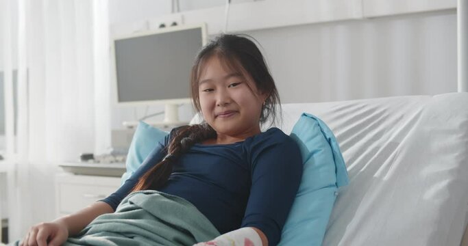 Smiling asian preteen girl lying in hospital bed with broken arm in colored cast