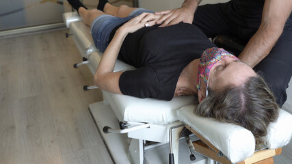 Physiotherapist with hands on hip and neck of woman lying down on stretcher with face mask on. Osteopath treating middle aged woman at office