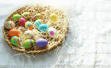 Fototapeta na wymiar On a white tablecloth there is a wicker plate with hay and colorful Easter eggs with patterns. Easter theme, Christian holiday