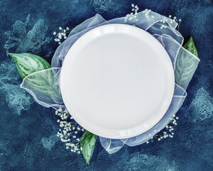 Spring season composition with a white plate and flowers on a blue background. Romantic, floral table setting with large leaves and gypsophila. Wedding Concept. Food mock up. Top view