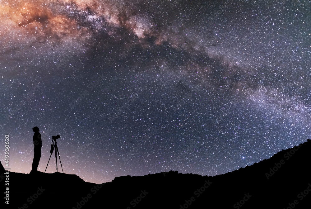 Wall mural the man silhouette with camera and tripod, stands on the hill and looking at the bright milky way ga