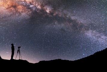 The man silhouette with camera and tripod, stands on the hill and looking at the bright milky way...