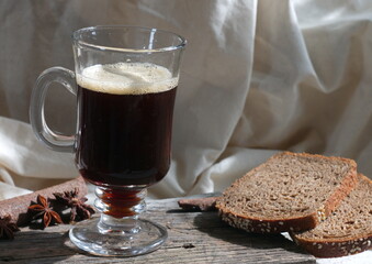 On a wooden table in a transparent glass, a dark drink with froth, and next to it anise and a slice of bread
