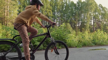 Tracking side-view full shot of sporty 12-year-old caucasian boy in helmet cycling on trail in park among trees on sunny day