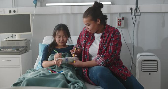 African adoptive mother and asian daughter painting hand gypsum together in hospital ward