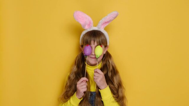 Portrait of funny cute little girl looks positively at camera, holds and covers eyes with two Easter colored eggs, being bunny, isolated on yellow background. Happy Easter and seasonal holiday concept