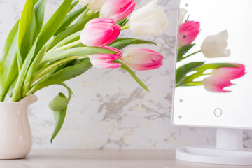 A bouquet of white and pink tulips with green leaves stands in a jug on the table and is reflected in the mirror. Selective focus.