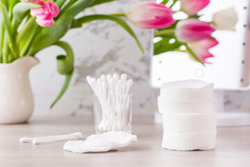 Fototapeta na wymiar Personal hygiene, cleanliness and skin care. Cotton pads and swabs in a glass on a table in front of a mirror and a bouquet of tulips in a vase