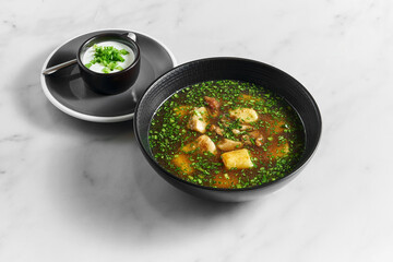 Soup, broth with white mushrooms with fresh herbs, sour cream and green onions in a black bowl on a light marble background