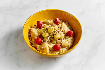 Tasty and healthy oatmeal with honey, apple, raspberry and sunflower seeds in a bright yellow bowl on a white marble background.