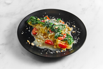 Pappardelle pasta with parsnip, cream sauce, blanched greens, tomatoes and nuts on a black plate on a white marble background
