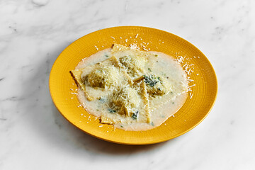 Tasty cooked ravioli with various fillings in a creamy sauce sprinkled with parmesan on a bright yellow plate on a white marble background