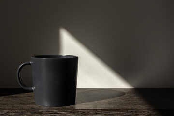 Black mug in bright sunlight on wooden table. Gray background with shadow and graphical triangular sun spot light. Minimal style mockup with copy space. Hard light and shadows concept.