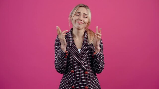 Concentrated young woman, who keeps her fingers crossed and makes a wish Beautiful mature young woman, blonde, in a pink jacket. Isolated on a pink background. Concept of life. People's emotions. 4k