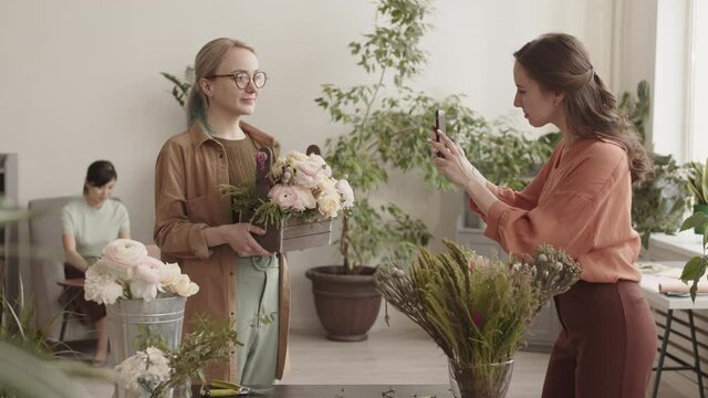 Medium long of young Caucasian woman taking picture of blond-haired female Colleague, holding wooden basket with flowers arrangement, standing in plant shop