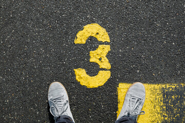 Men with grey sneker and jeans stand on the number 3, sign on the asphalt, yellow painted number...