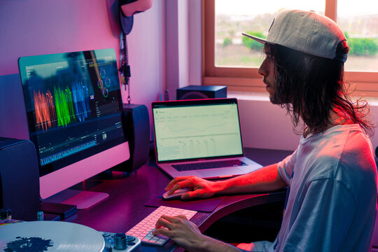 Creador de contenido on line con gorra, sentado delante de su ordenador de sobremesa y su ordenador port&aacute;til, con un programa de edici&oacute;n de color de v&iacute;deo y las estad&iacute;sticas de su canal en el port&aacute;til