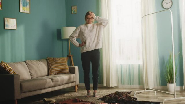 The girl is standing in the middle of a spacious room next to the sofa, women's clothes are scattered on the floor. The lady leaves the building with a dissatisfied look