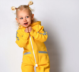 Playful toddler girl with two ponytails on her head is dressed in a warm yellow sports jumpsuit. Child with an interested facial expression looks away on a gray background. Place for text.