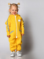 Small cute blonde smiling baby girl in yellow warm comfortable jumpsuit standing and looking aside over grey wall background. Happy childhood and trendy children wear concept