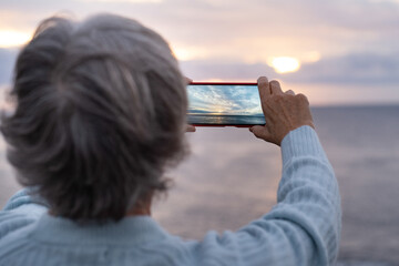 Tourist takes a picture to the sunset over the sea - back view of a gray haired woman