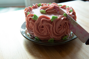 A strawberry buttercream decorated double layer chocolate cake. The icing is pink rosettes with green leaves. The thick, silky, creamy and smooth berry flavour celebration cake has a knife cutting it.