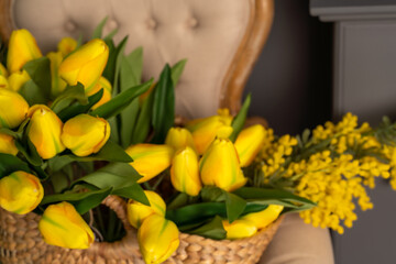 A large bouquet of yellow tulips and a yellow mimosa in a wicker basket. The basket sits on a beige chair against a gray wall.