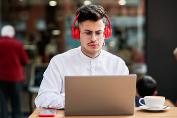 Fototapeta premium young man sitting on the terrace of a coffee shop with a laptop and red headphones while drinking a coffee.