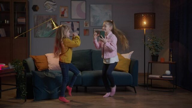 Cute little American girls dance in living room, watching TV and recoding video for instagram. Concept of happiness life and stay at home during covid virus pandemic around the world.Evning night time