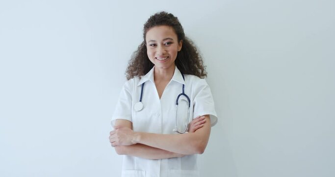 Portrait of satisfied african american female doctor wear medical coat crossing arms looking at camera and smiles standing by white wall. Confident mixed race woman, young curly nurse