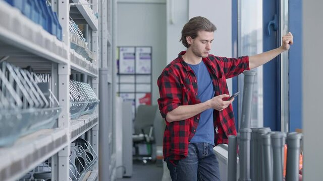 Absorbed Caucasian young man surfing Internet on smartphone standing between rows in hardware store. Engrossed handsome millennial using social media online buying goods.