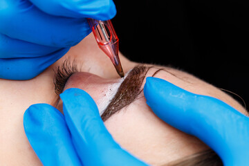 Beautician tattooing a woman's eyebrows using special equipment during permanent make-up, close-up.