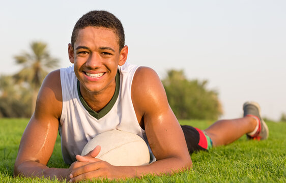Happy fit black teen athlete resting on the grass with a rugby ball. Sports lifestyle.