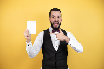 Young man with beard wearing bow tie and vest smiling and showing blank notebook