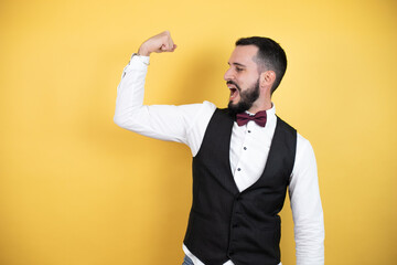 Young man with beard wearing bow tie and vest showing arms muscles smiling proud