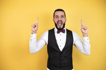 Young man with beard wearing bow tie and vest amazed and surprised looking at the camera and pointing up with fingers and raised arms
