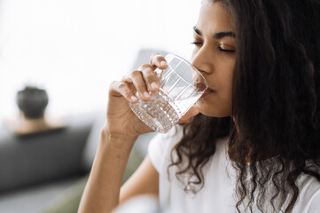 Healthy lifestyle. African american young woman drinks a glass of water while sitting on the sofa at home. Beautiful healthy woman follow healthy lifestyle