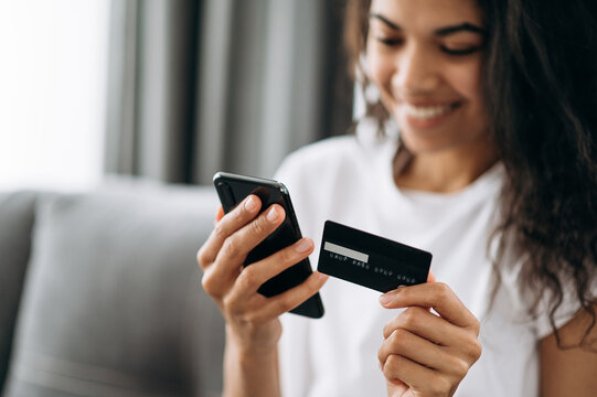 A credit card and smartphone in female hands. Joyful african american young woman in defocus looks at the phone screen, shopping online. Credit card and phone in the focus