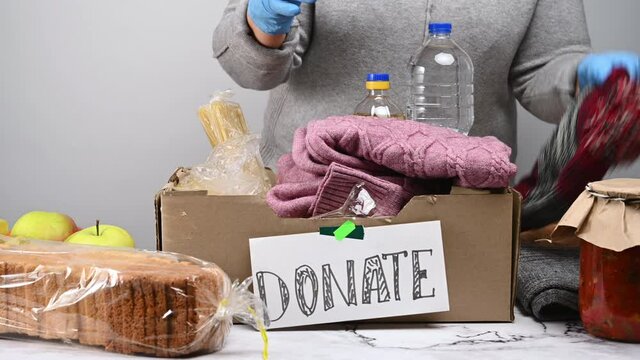 woman in a gray sweater puts various things and food in a box with the inscription donate. Helping the poor concept, volunteering 