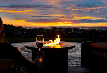 A woman drinks a glass of wine in front of a fire pit on the luxury patio of a hillside home overlooking city lights. - obrazy, fototapety, plakaty