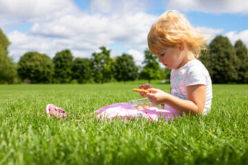 Little girl sitting on the grass in the meadow and having snack on summer day.