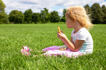 Little girl sitting on the grass in the meadow and having snack on summer day.