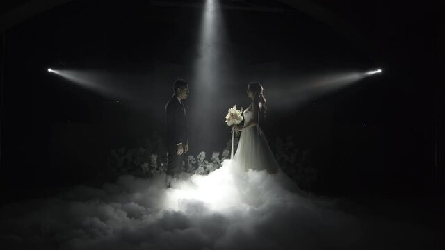 A lovely couple, makes a step to the meeting, on the floor a lot of white smoke, the spotlight shines right on them and behind the black background.